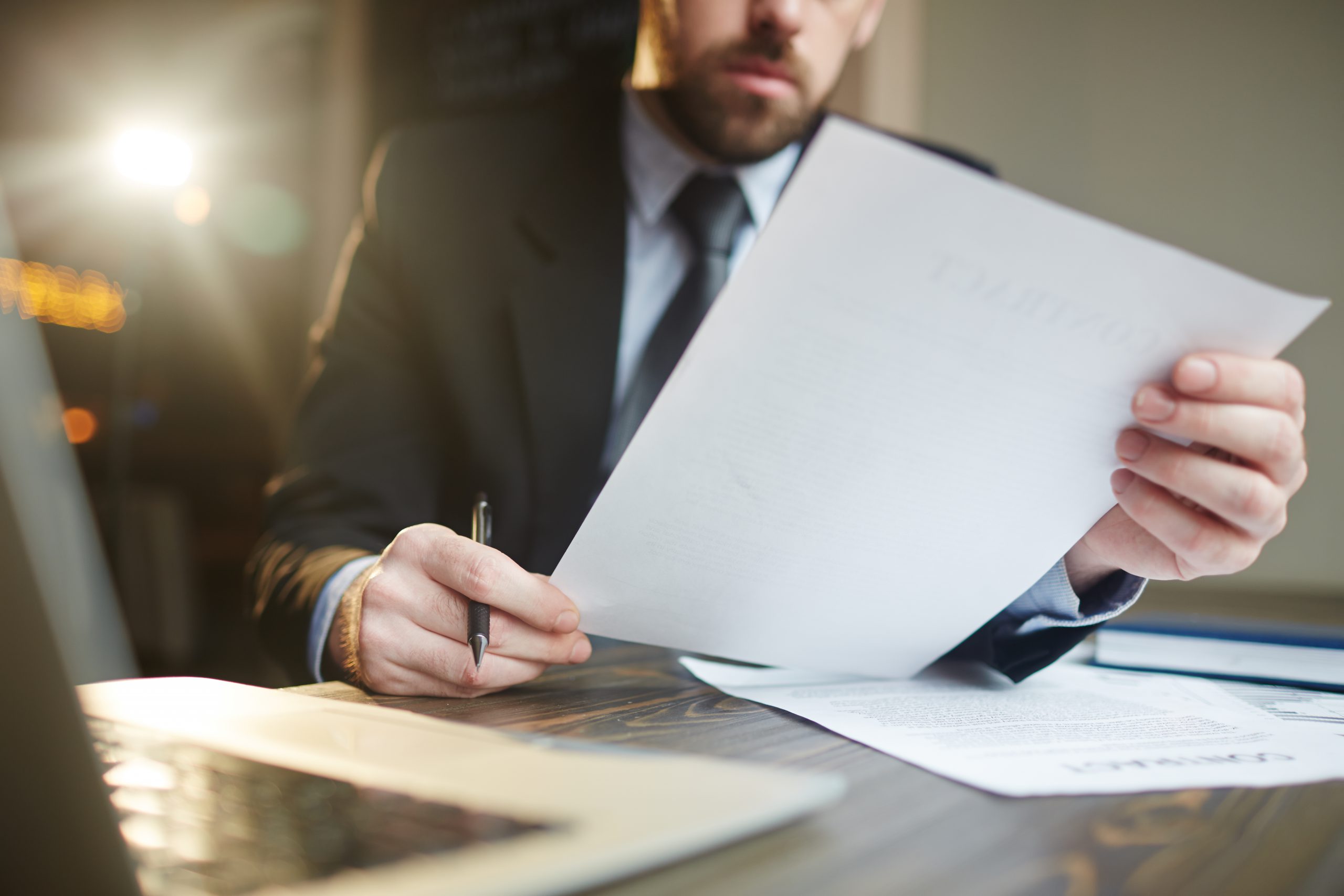 Portrait of modern bearded businessman holding papers in hands, reading and analyzing contract documentation at desk with laptop